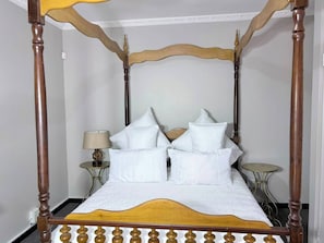 A four-poster bed with white bedding and multiple pillows, a bedside table with a lamp, and a small round table.