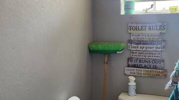 A small bathroom with a toilet, a broom, a roll of toilet paper, and a sign with toilet rules.