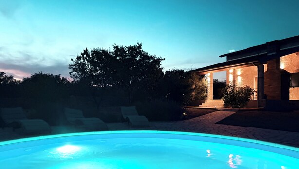 Une piscine à bord bleu clair, un espace extérieur couvert avec des piliers en pierre, et un bâtiment à l’éclairage chaleureux.
