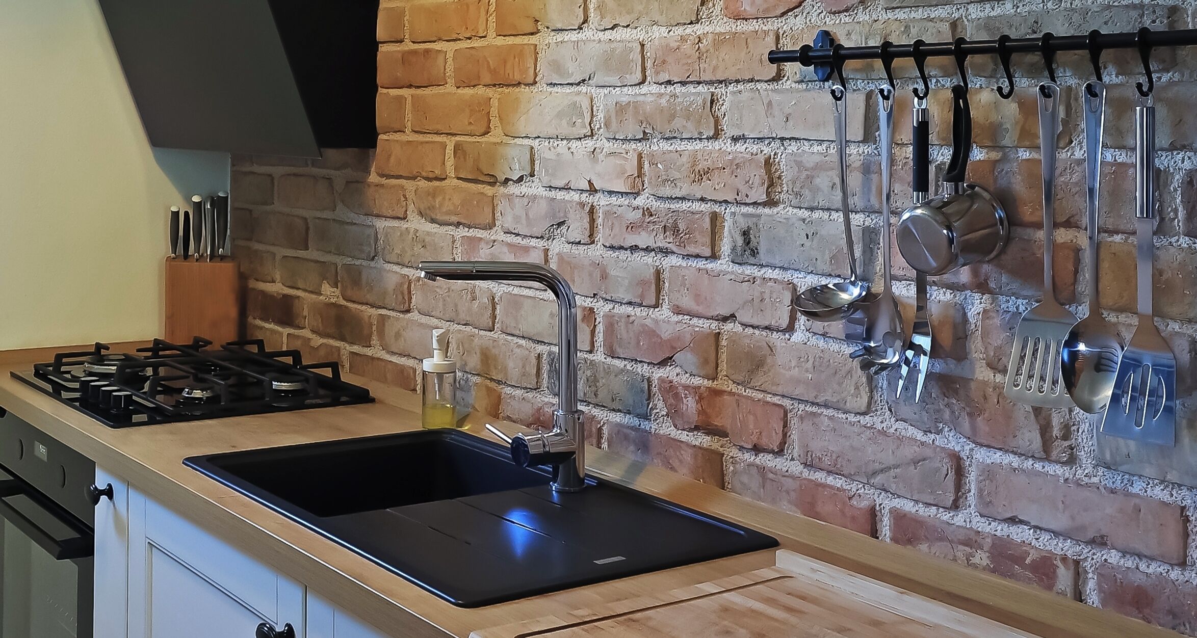 Cucina con piano cottura a gas, lavello nero e utensili appesi a una parete in mattoni.