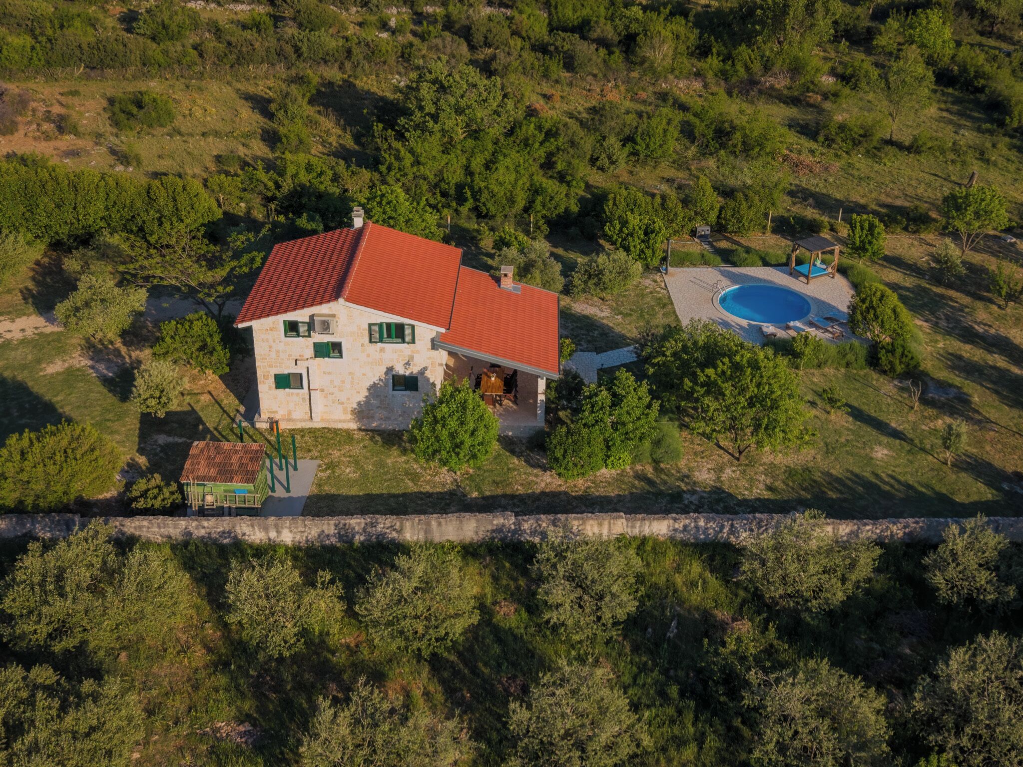 Vista aérea de una casa con techo rojo, piscina y un muro de piedra en primer plano.