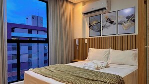 Una camera d'albergo con un'ampia finestra, un letto con coperta color crema, una testiera in legno e tre opere d'arte appese al muro.