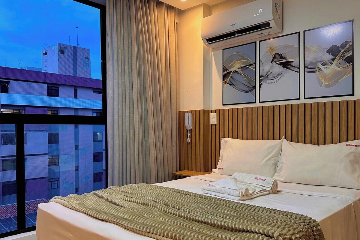 Ein Hotelzimmer mit großem Fenster, einem Bett mit beiger Decke, einem Holzkopfschild und drei Wandbildern.