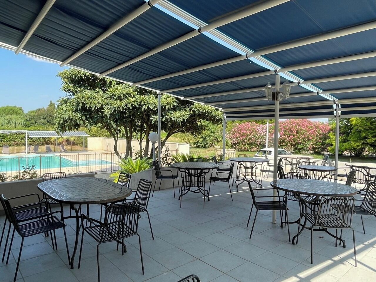 An outdoor patio with tables and chairs, a covered area, and a pool in the background.