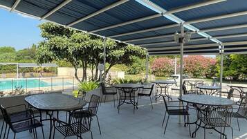 Um pátio externo com mesas e cadeiras, uma área coberta e uma piscina ao fundo.