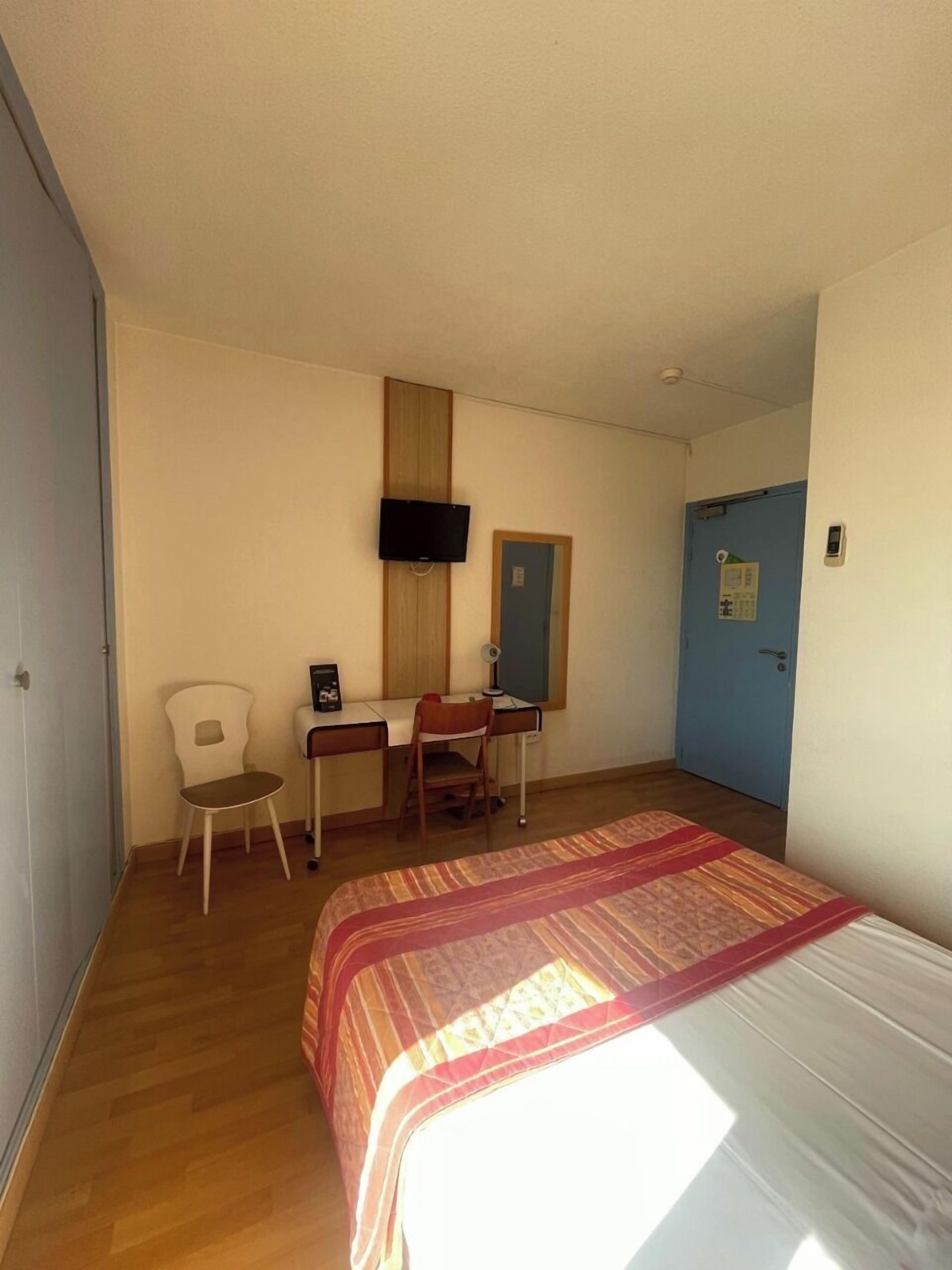 Una habitación con una cama, un escritorio, una silla, un televisor montado en la pared y una puerta.