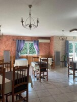 Un comedor con sillas y mesas de madera, una araña y ventanas amplias con cortinas.