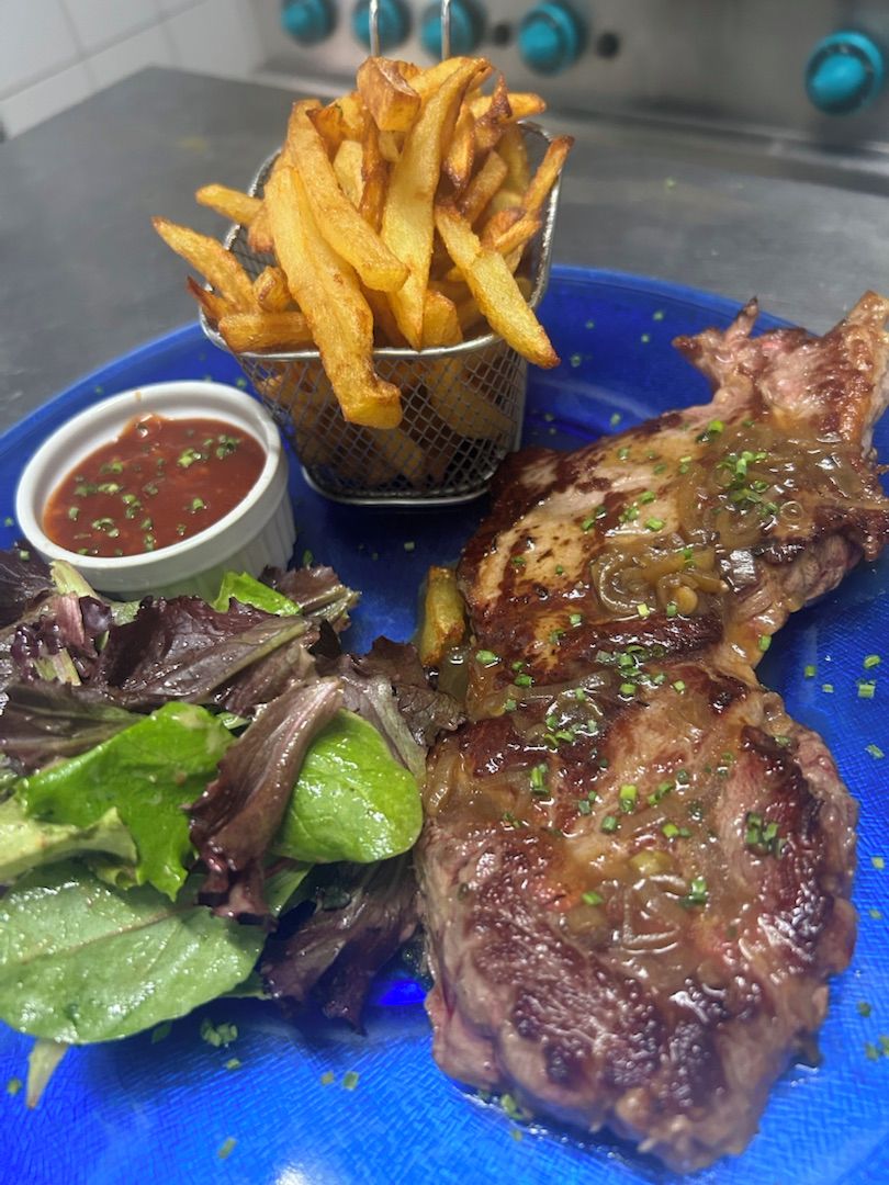 Een bord met een gegrilde steak, frietjes, een klein kommetje saus en een salade.