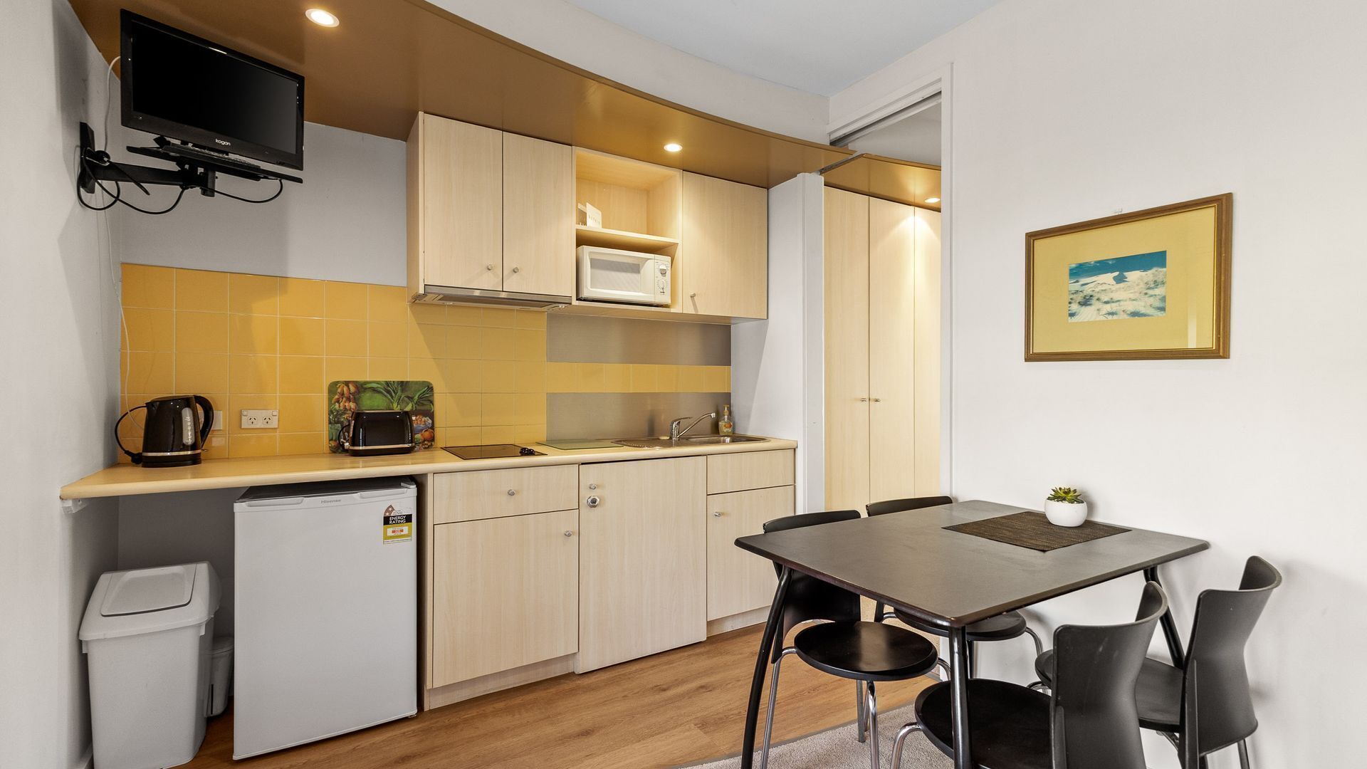 A compact kitchen with a microwave, toaster, and coffee maker on the counter, a small refrigerator, and a dining table with chairs.