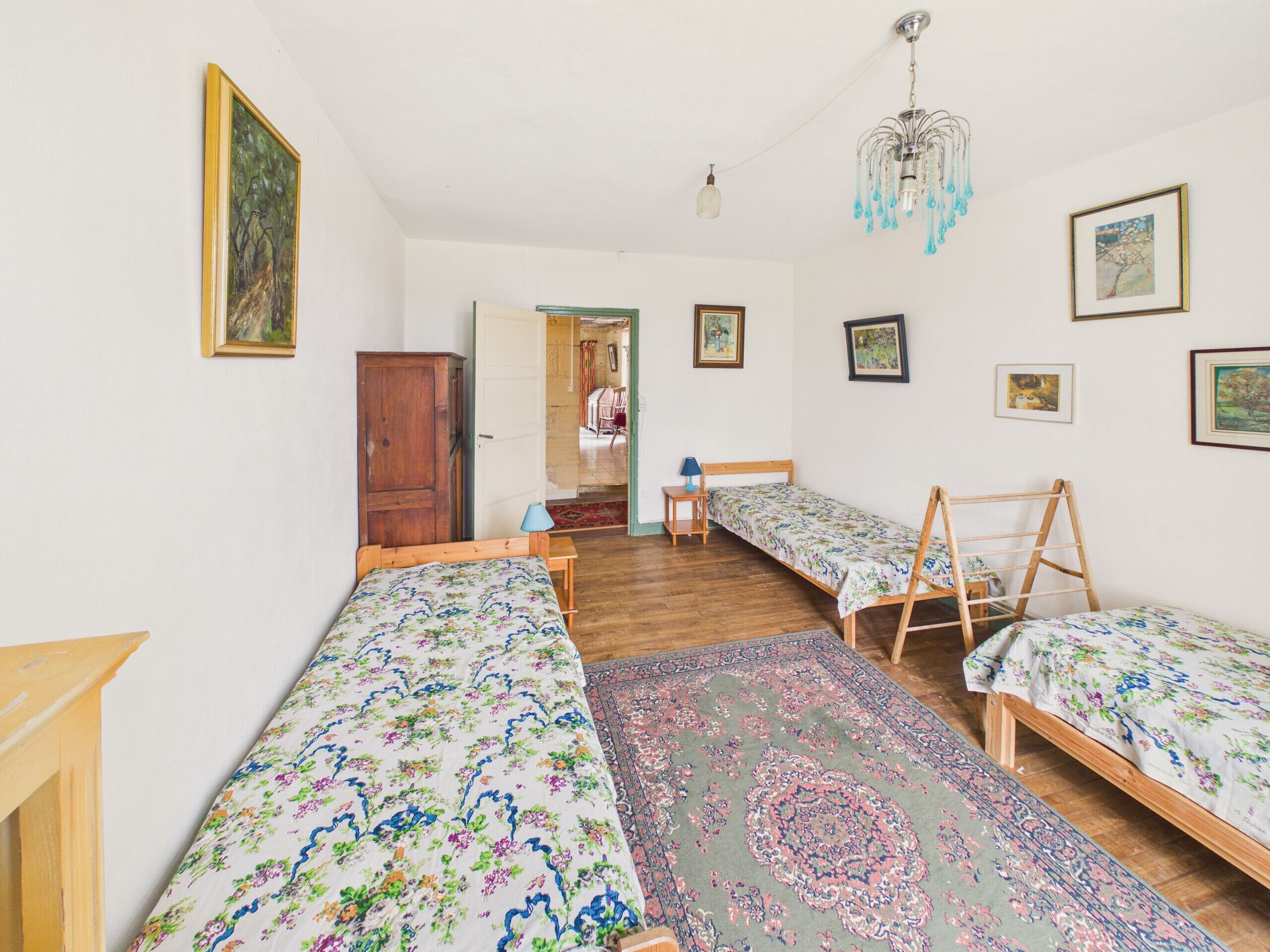 Una habitación con tres camas individuales, un armario de madera, una lámpara de araña y varias obras de arte enmarcadas en las paredes.