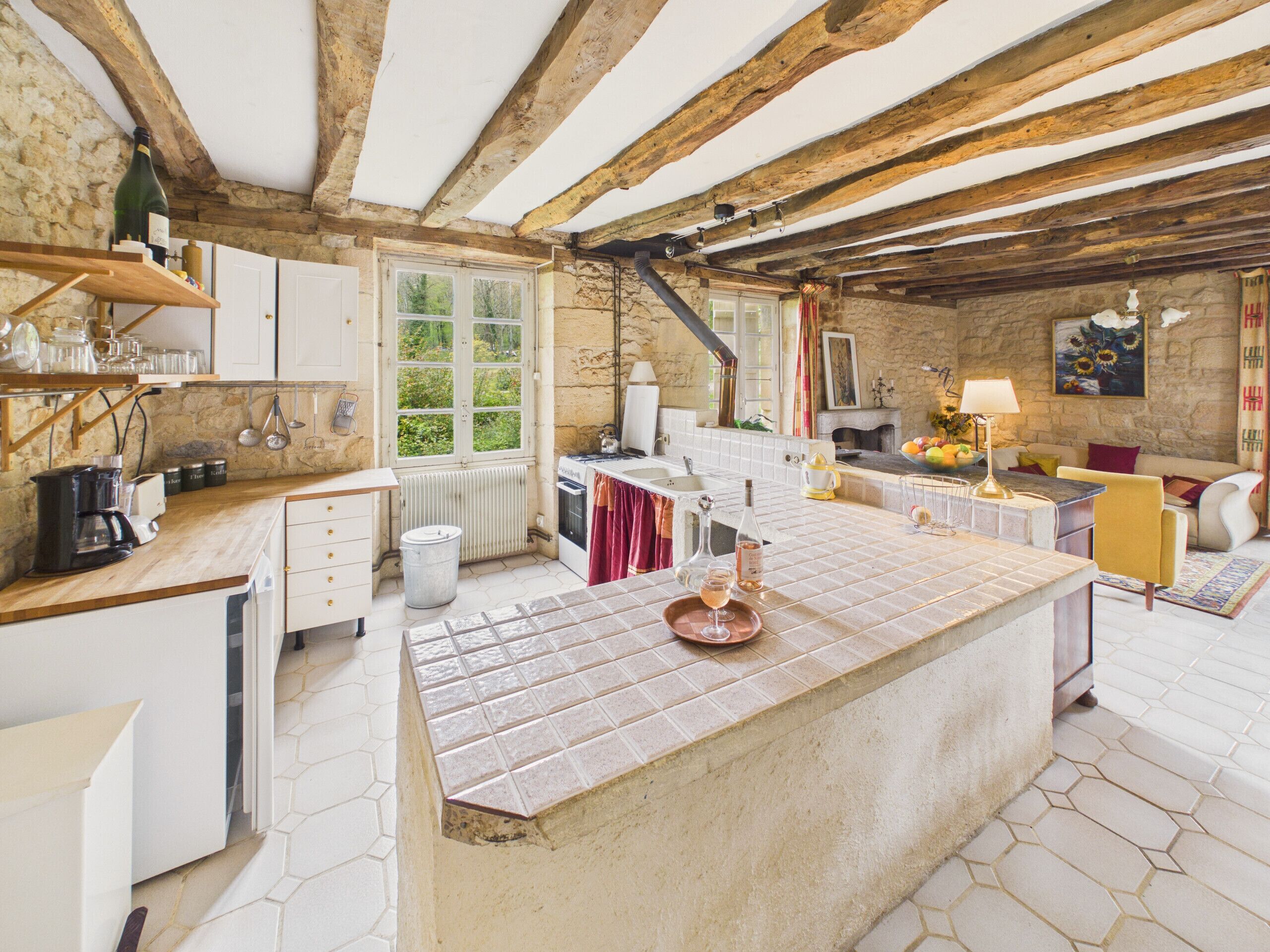 A rustic kitchen with a large island, wooden beams, stone walls, and a view of greenery outside.