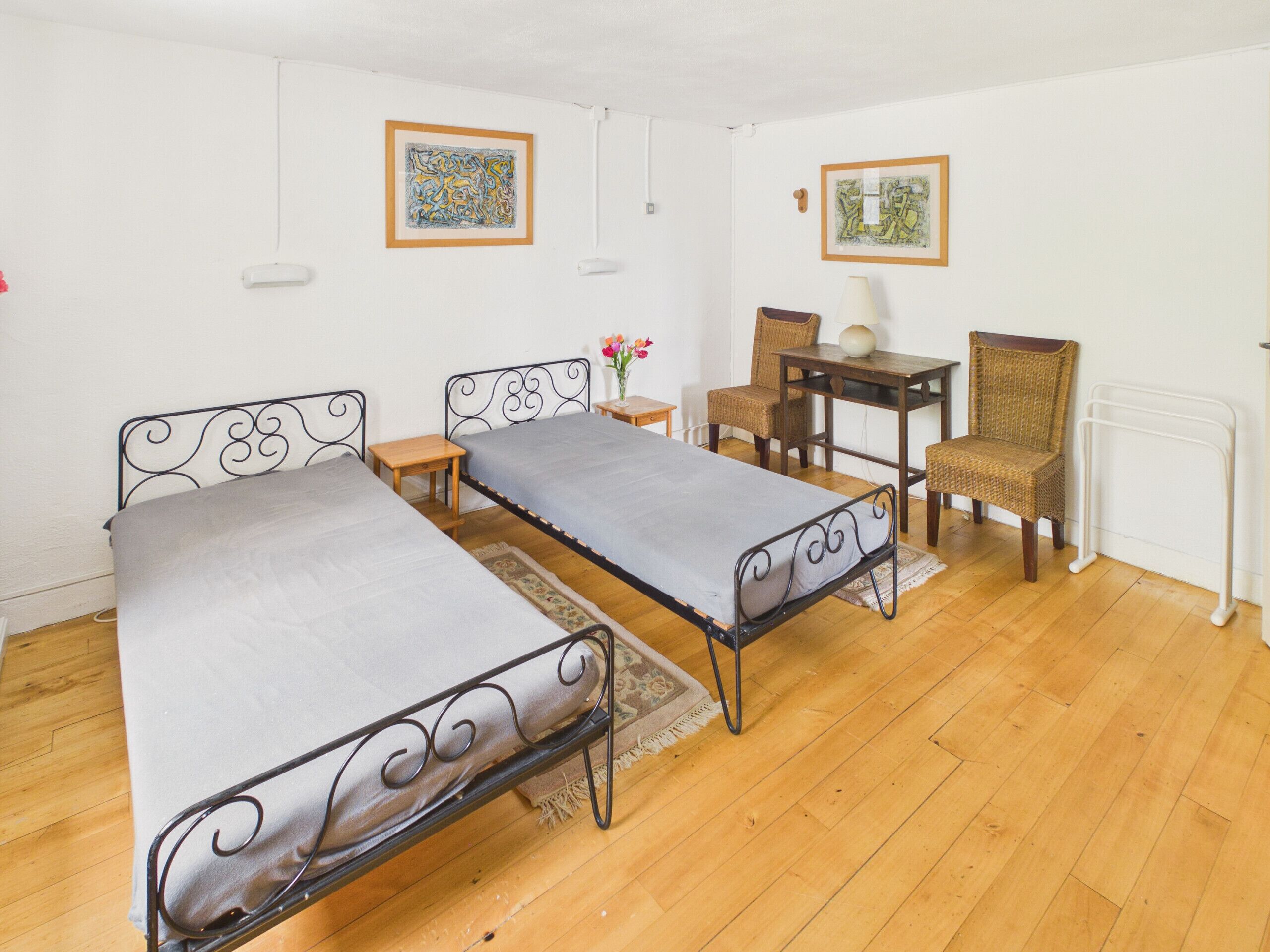 Una habitación con dos camas de metal, un escritorio de madera, sillas de mimbre y dos cuadros en las paredes.