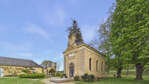Een klein stenen gebouw met een klokkentoren, omringd door een verzorgd gazon en bomen.