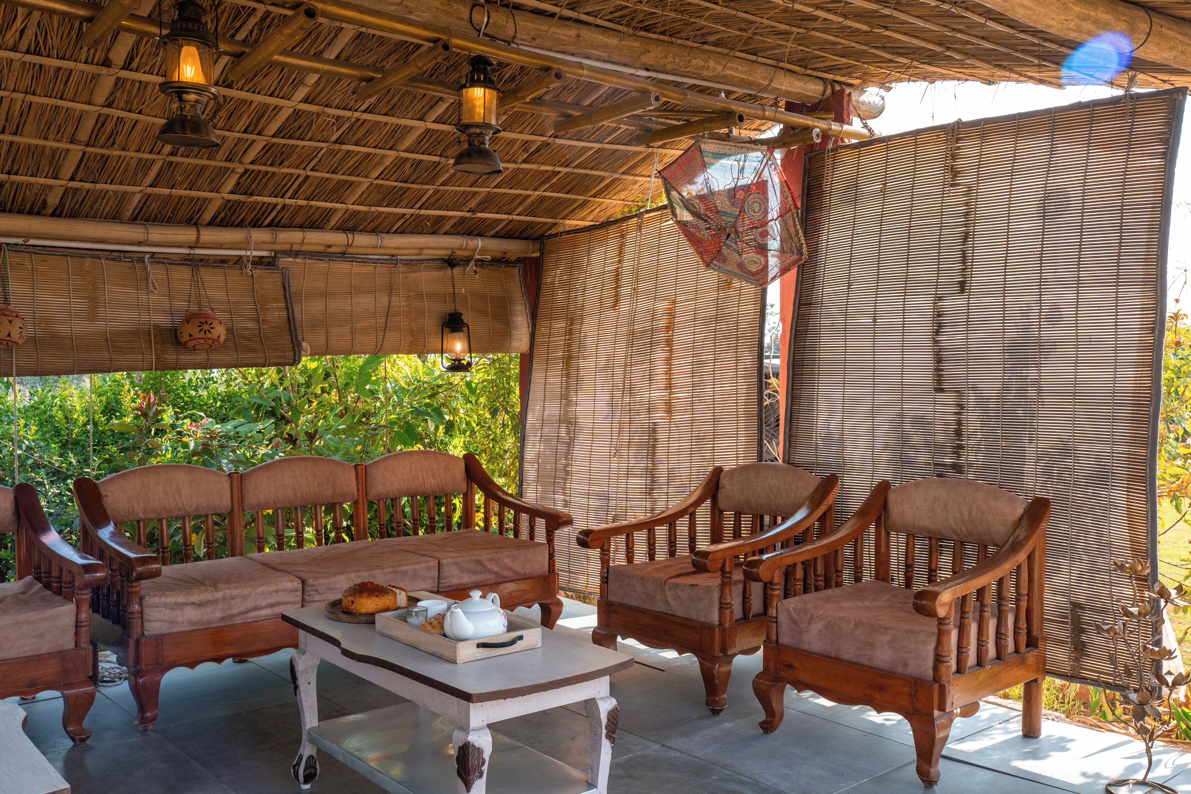 A covered outdoor seating area with wooden furniture, a table with a teapot and tray, and bamboo blinds.