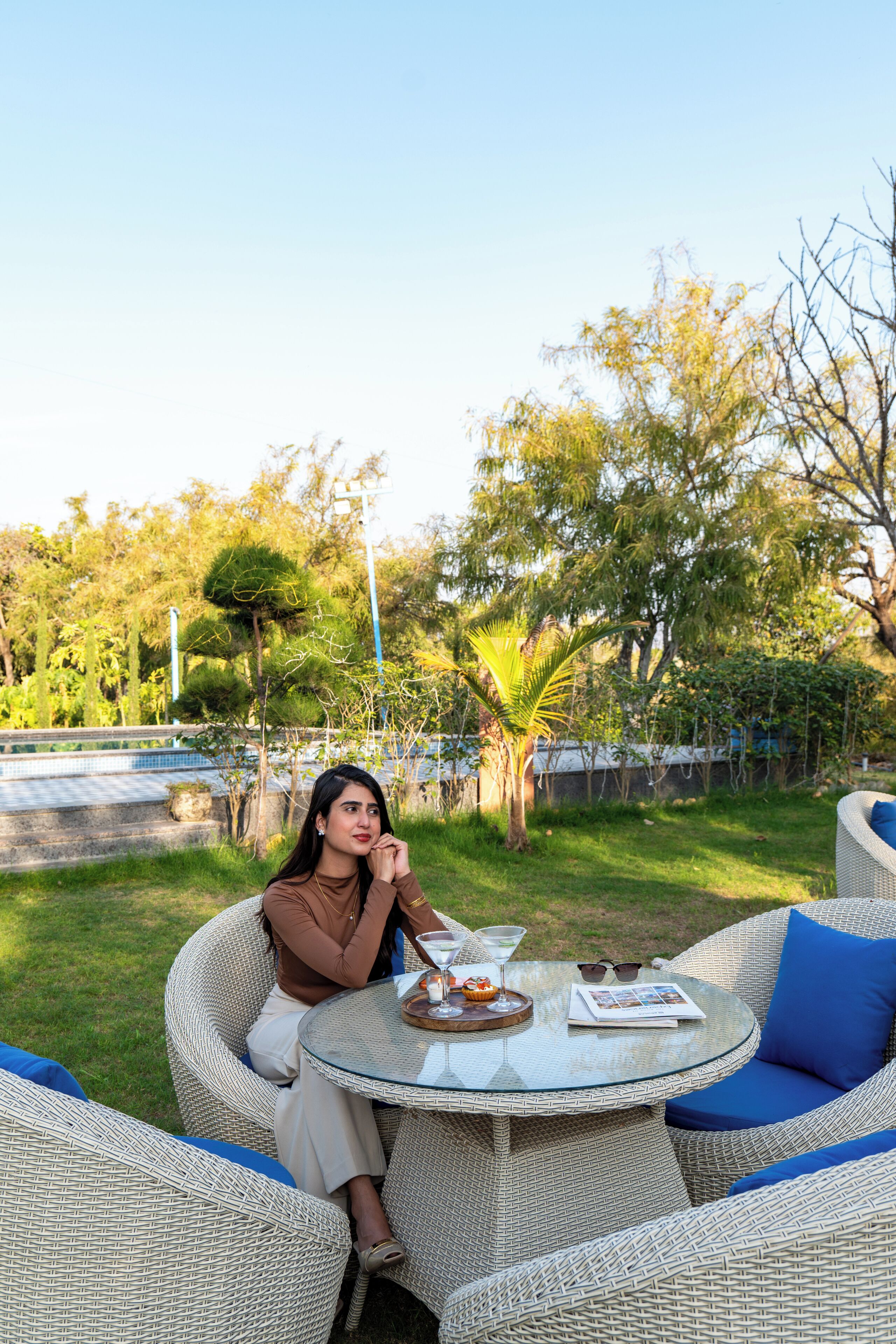 A woman sitting on wicker outdoor furniture with a glass table, surrounded by greenery and trees.