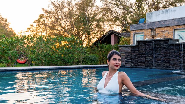 A swimming pool with clear water, surrounded by greenery and a stone wall. - Krishna Serenity Petfriendly 7BHK Farm (Peesangan)
