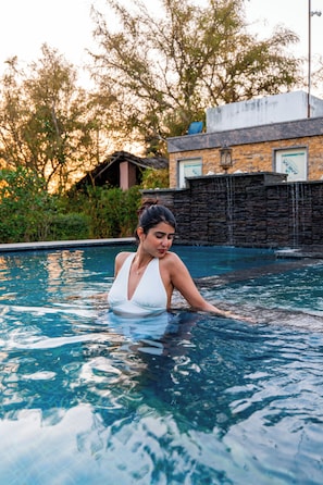 Une femme vêtue d’un maillot de bain blanc se trouve dans une piscine aux eaux cristallines, avec un mur de pierre et de la verdure en arrière-plan.
