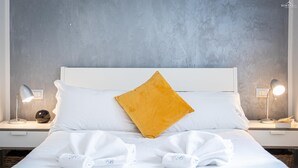 Een net opgemaakt bed met witte lakens en één geel kussen, twee nachtlampjes en een gestructureerde grijze muur.