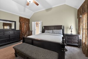 A bedroom with a large bed, a dresser, a nightstand, a bench, a ceiling fan, and a door leading to another room. - Spacious Lakefront Home | Memphis Retreat (Memphis)