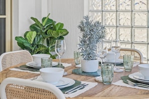 Une table à manger dressée avec de l’assiette blanche, des bols et de la verrerie transparente, entourée de chaises en osier.