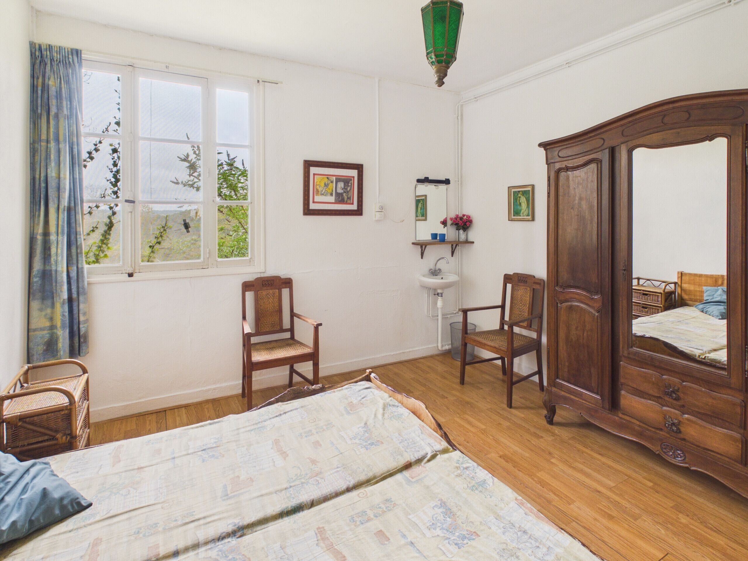 Une pièce comprenant un lit, deux chaises, une armoire en bois, une fenêtre avec des rideaux, un lavabo fixé au mur et des tableaux encadrés.