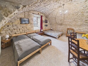 Une chambre rustique avec un plafond en pierre, un lit en bois, une petite table avec une chaise et une fenêtre ornée d’un rideau rouge.