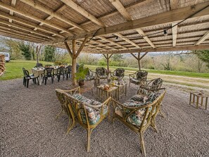 Une terrasse extérieure couverte, aménagée avec des meubles en osier, une table et des chaises, et offrant une vue sur la campagne.