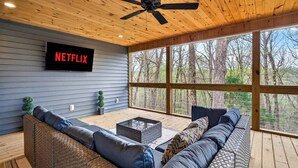 Ein hölzerner Deckenventilator, ein Netflix-Schild, ein großes Fenster mit Blick auf Bäume und ein Sofa mit Kissen.