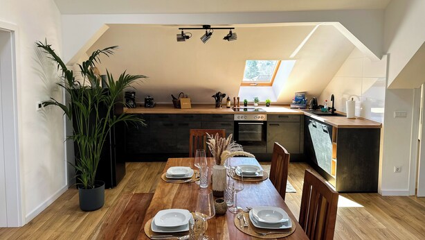 Zona pranzo con tavolo in legno apparecchiata per quattro persone, cucina con lucernario e una pianta in vaso.