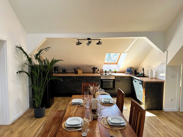 Comedor con mesa de madera dispuesta para cuatro, cocina con tragaluz y una planta en maceta.