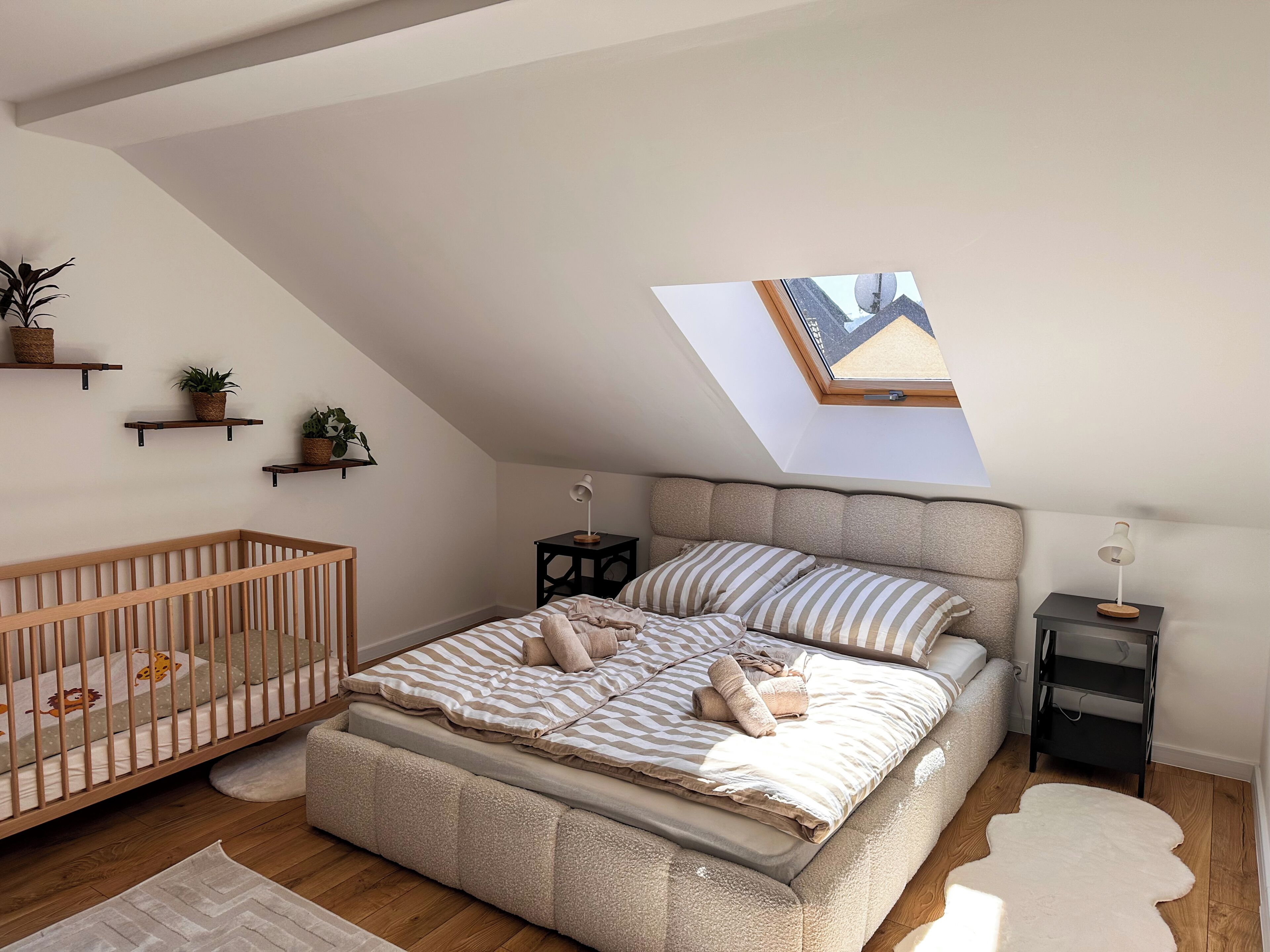 A bedroom with a sloped ceiling, a skylight, a crib, a bed with striped bedding, a bedside table, and a nightstand.