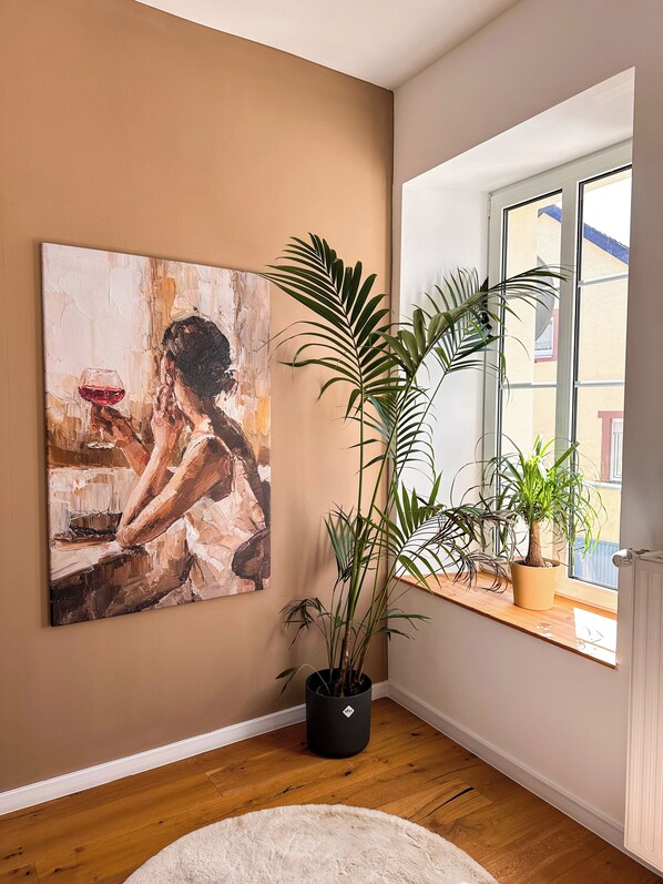 A room with a large abstract painting, a potted plant, a window with a view, and a wooden floor.