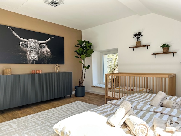 Un dormitorio moderno con una cama grande, una cuna, una consola gris, un televisor montado en la pared y una planta en maceta.
