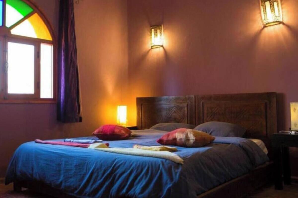 A bedroom with a bed, pillows, a nightstand, a window with curtains, and wall-mounted lights.