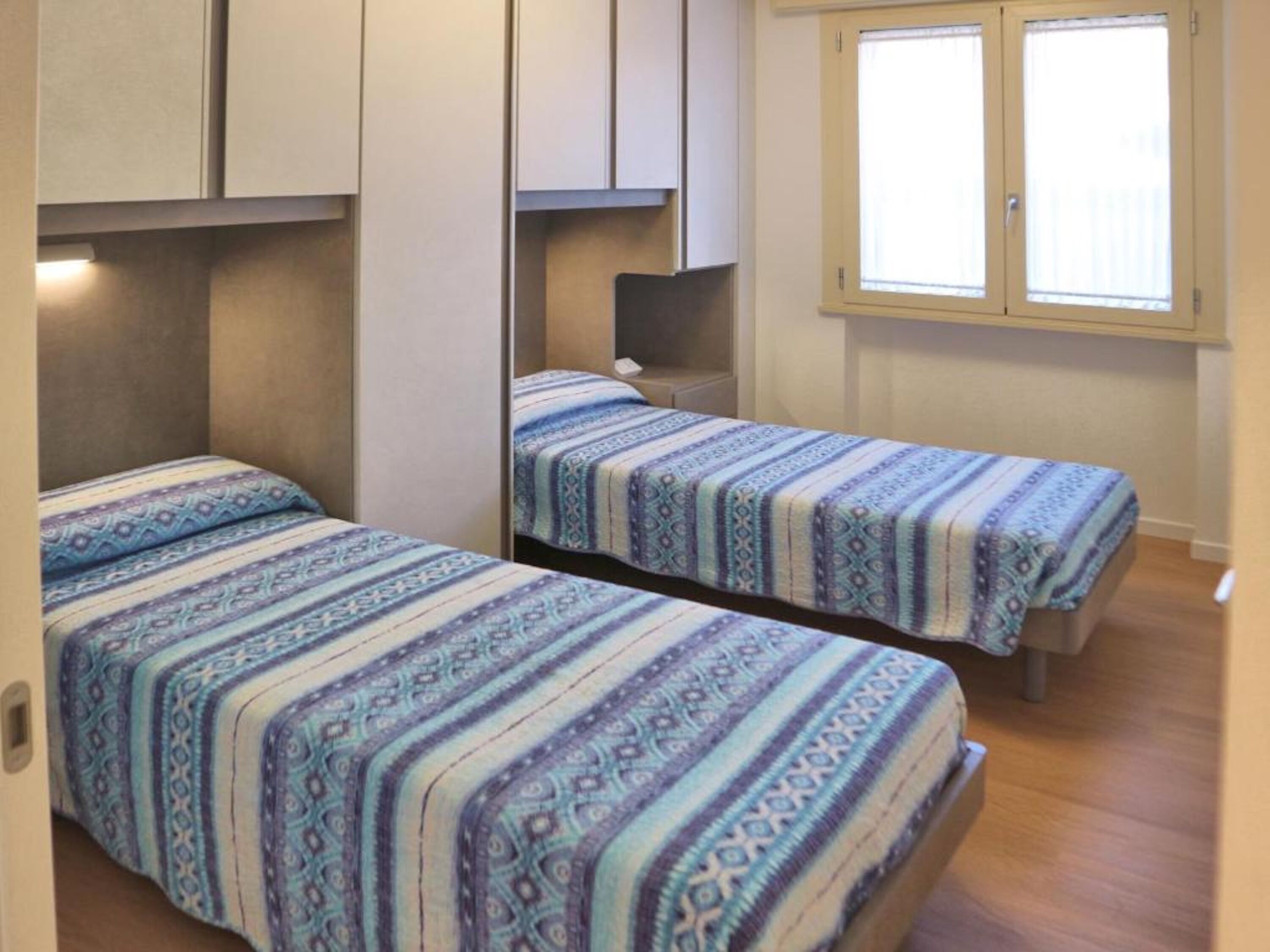 A room with two beds, each with patterned bedding, a window with blinds, and a wardrobe.