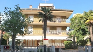 A multi-story building with balconies, a palm tree, and a gated entrance. - Modern Central Flat With two Balconies (Lignano Sabbiadoro)