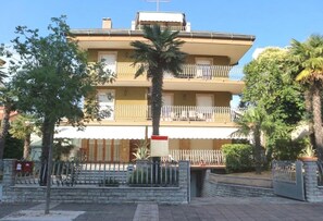 A multi-story building with balconies, a palm tree, and a gated entrance. - Modern Central Flat With two Balconies (Lignano Sabbiadoro)