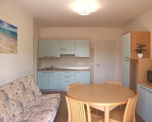 A compact living space with a kitchenette, a dining table with chairs, a sofa, and a ceiling light fixture. - Modern Central Flat With two Balconies (Lignano Sabbiadoro)