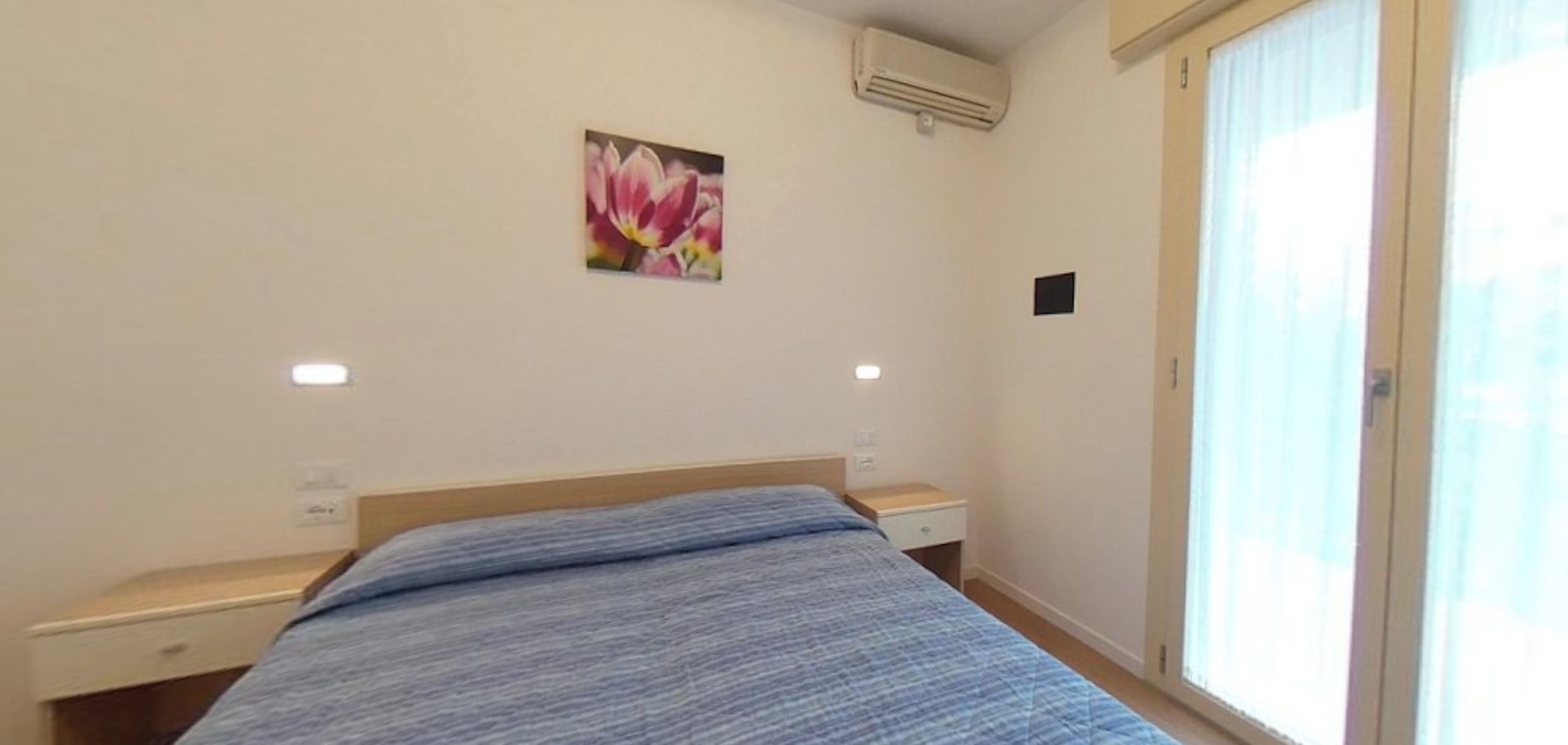 A bedroom with a bed, a nightstand, a wall-mounted air conditioner, and a window with blinds.