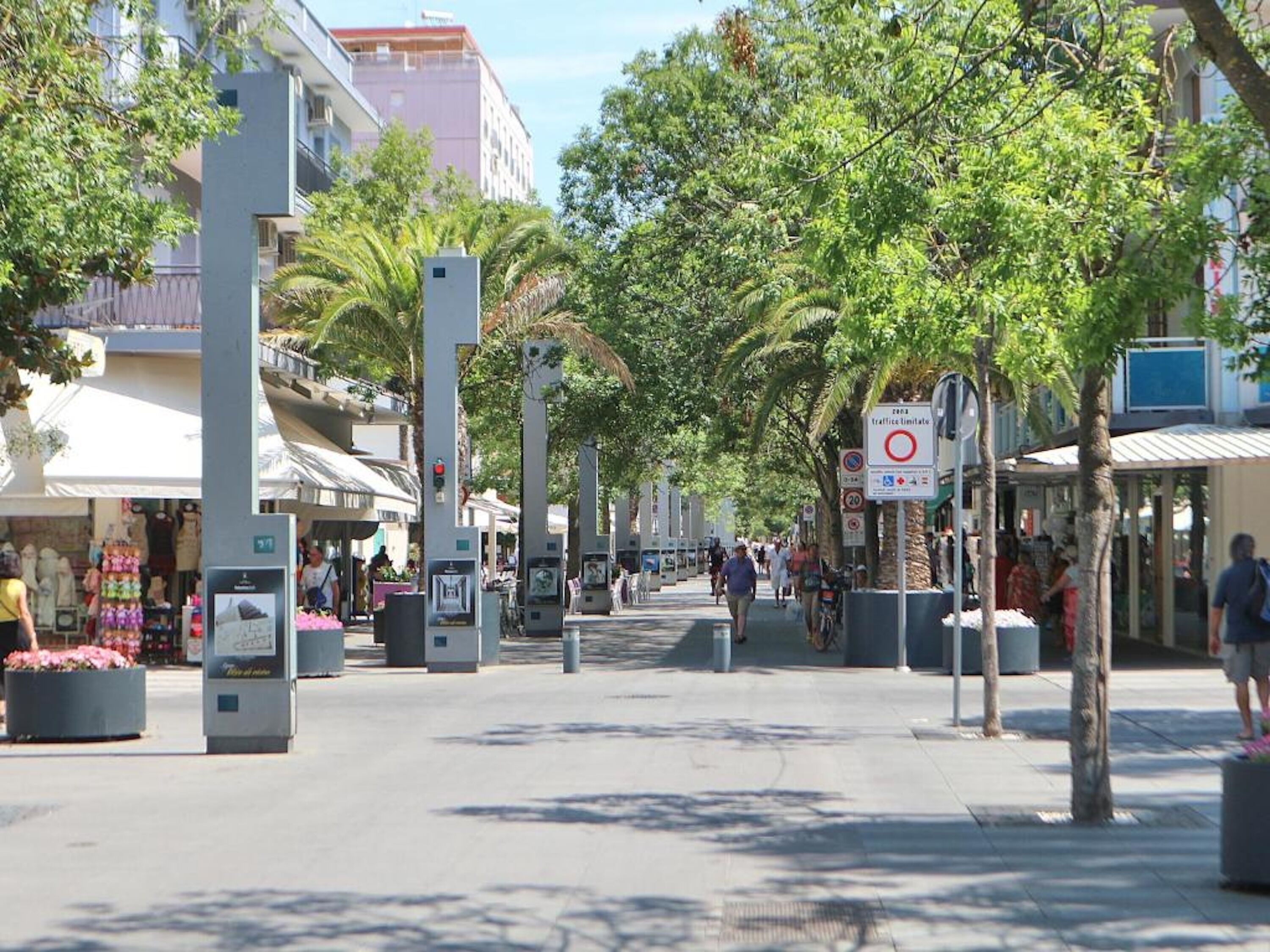 A pedestrian street lined with trees and buildings, with a traffic limit sign visible.
