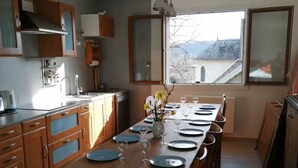 A kitchen with a long dining table set for six, wooden cabinets, a window with a view, and a hanging light fixture. - GÏTE DE LA CHAPELLE (Talissieu)