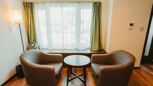 Et værelse med trægulv, to brune lænestole, et lille rundt bord, et vindue med grønne gardiner, en gulvventilator og en vægmonteret lampe.