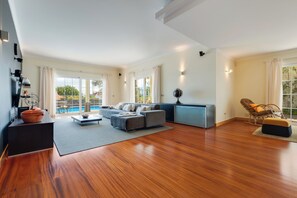 Amplia sala de estar con sofá, mesa de centro y televisor. Cuenta con ventanas grandes y vista a una piscina.