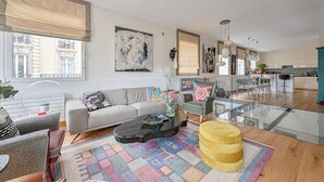 Uma sala de estar moderna com sofá, mesa de centro e um banquinho, uma área de jantar com cadeiras e mesa, e uma cozinha com armários e bancada.