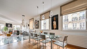 Quarto espaçoso com mesa de jantar de vidro, cadeiras modernas e grandes janelas com vista para edifícios.