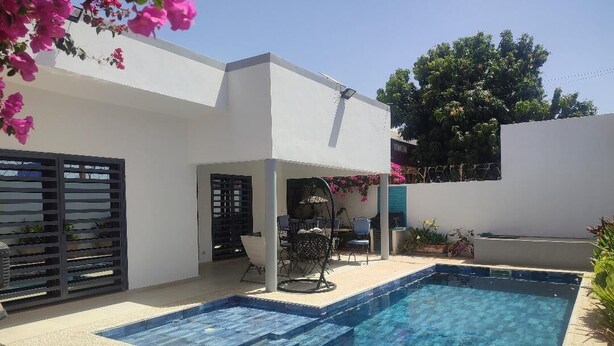 A swimming pool with two separate sections, a covered seating area with chairs, and a white building with a flat roof.