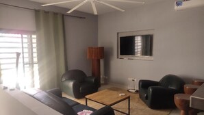 A living room with a ceiling fan, a flat-screen TV mounted on the wall, a sofa, two armchairs, a coffee table, and a lamp.