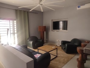 A living room with a ceiling fan, a flat-screen TV mounted on the wall, a sofa, two armchairs, a coffee table, and a lamp.