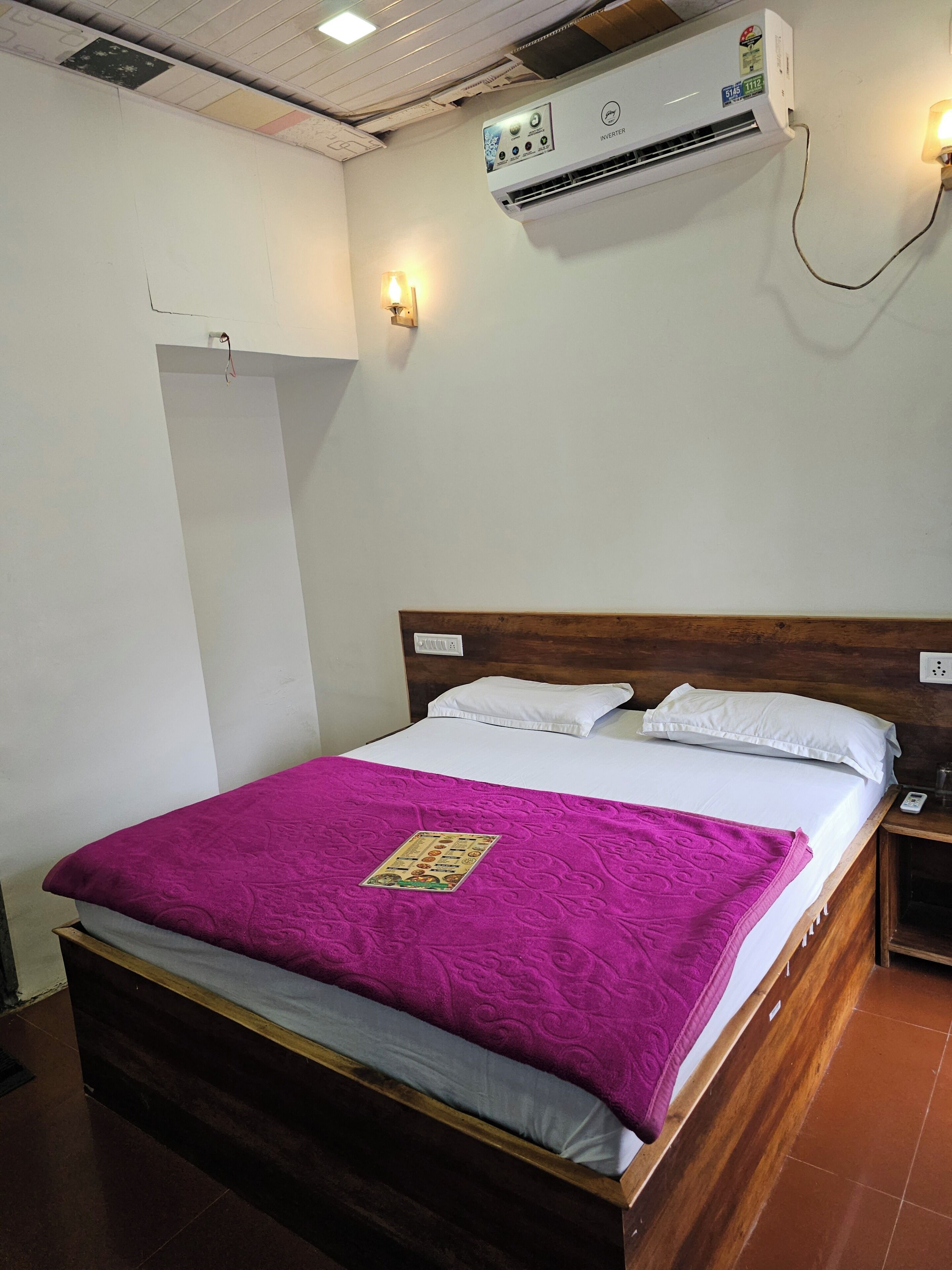 A hotel room with a bed, a nightstand, an air conditioning unit, and a wall-mounted light fixture.