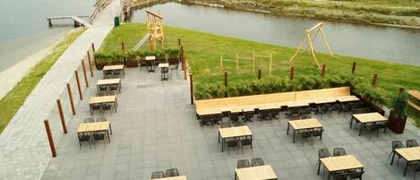 A riverside outdoor seating area with tables and chairs, a wooden swing set, and a pathway leading to a dock.